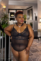 Model wearing plus size black lace bodysuit standing by staircase in upscale living room setting